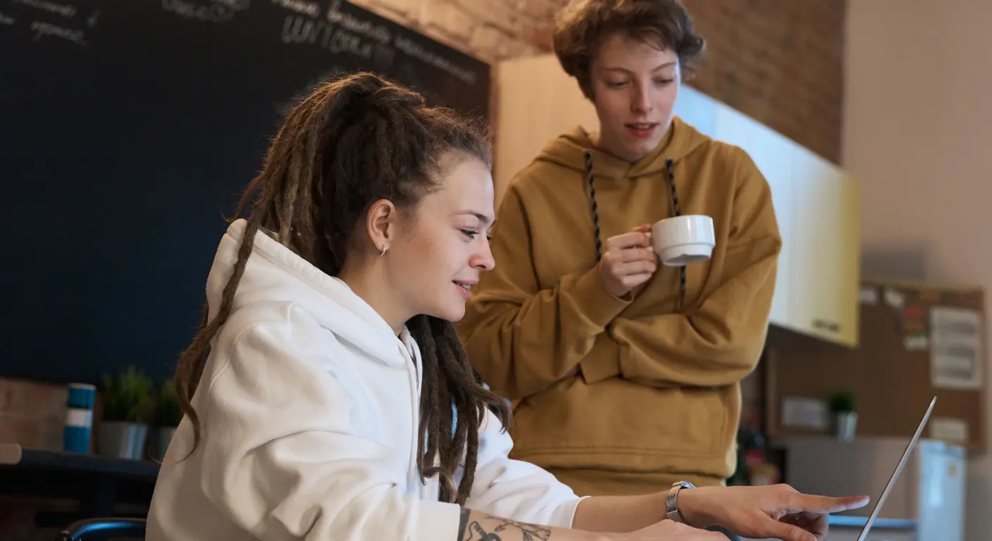 Two people working together at a laptop in a relaxed, informal office — one seated and typing while the other stands beside her with a coffee mug, looking at the screen